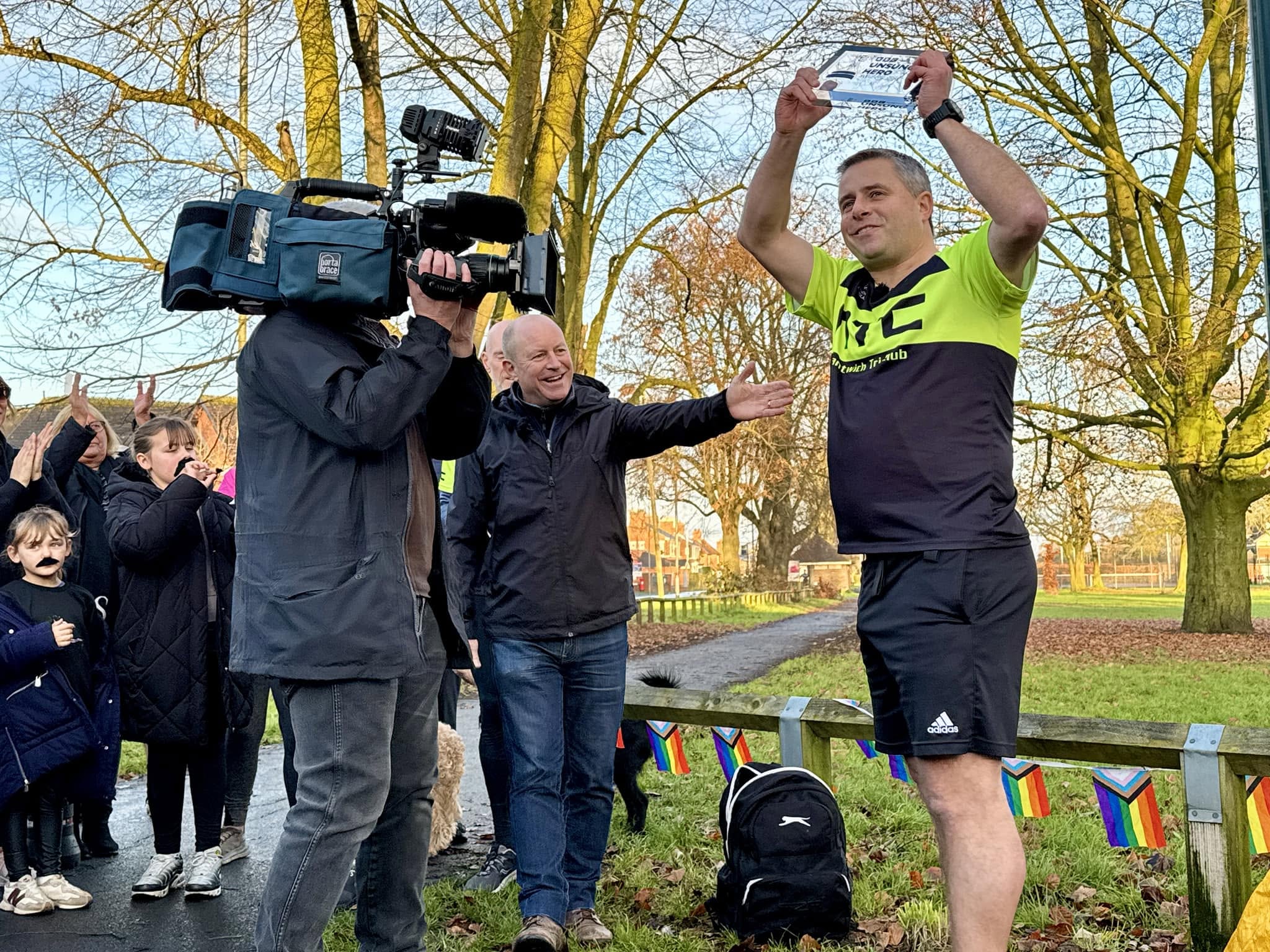 A television camera films a community sports volunteer receiving recognition in a park